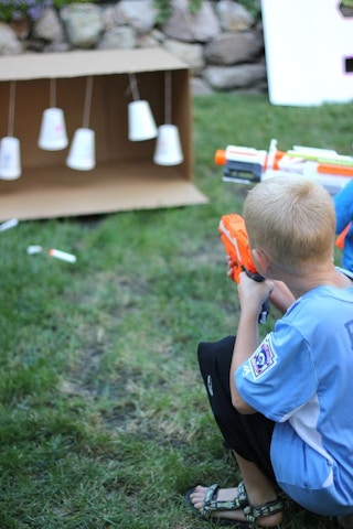 Two children shooting Nerf guns at some cups dangling from a DIY Nerf range made of cardboard and plastic cups.