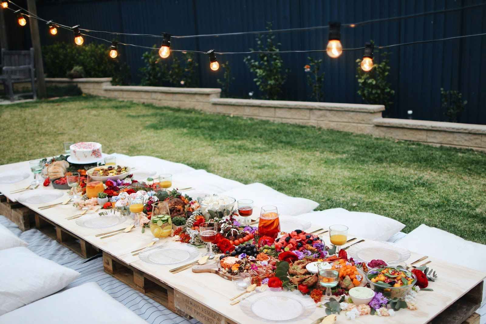 Several wooden pallets laid out in a backyard as a picnic table, with a table cloth, table settings, and flowers decorating it, and pillows for seats around them.