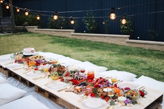Several wooden pallets laid out in a backyard as a picnic table, with a table cloth, table settings, and flowers decorating it, and pillows for seats around them.