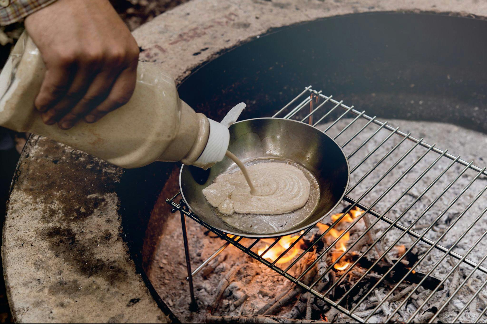 Someone squirting pancake batter from an old ketchup bottle into a pan over a campfire