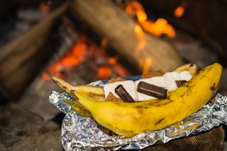 A banana cut in half, with chocolate and marshmallows inside, sitting on aluminum foil next to a campfire