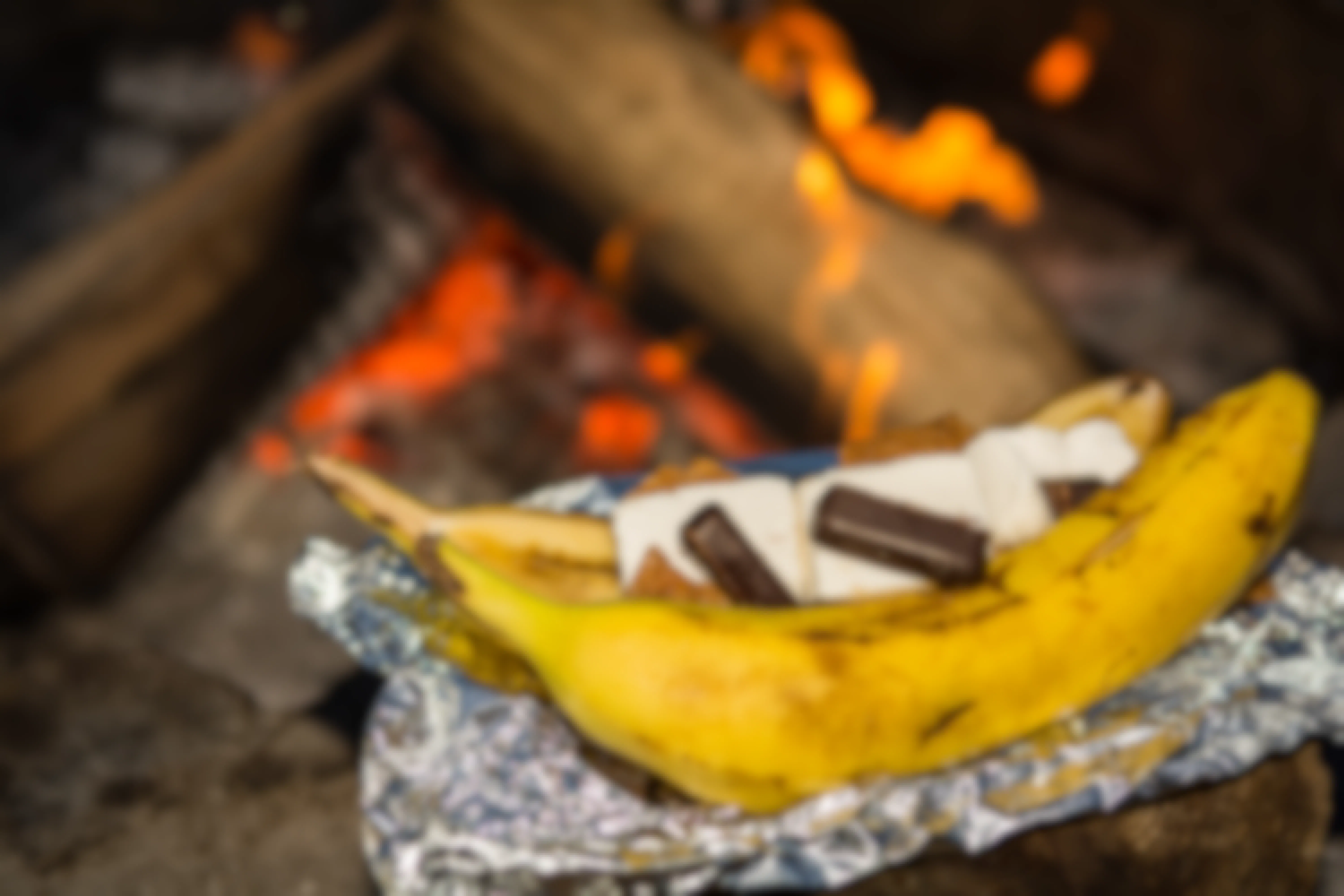 A banana cut in half, with chocolate and marshmallows inside, sitting on aluminum foil next to a campfire