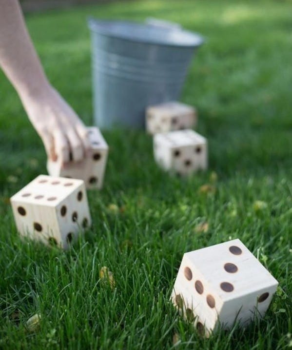 A person's hand reaching down to pick up a giant DIY dice block that is on the lawn next to more DIY dice and a metal bucket.