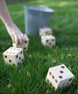 A person's hand reaching down to pick up a giant DIY dice block that is on the lawn next to more DIY dice and a metal bucket.