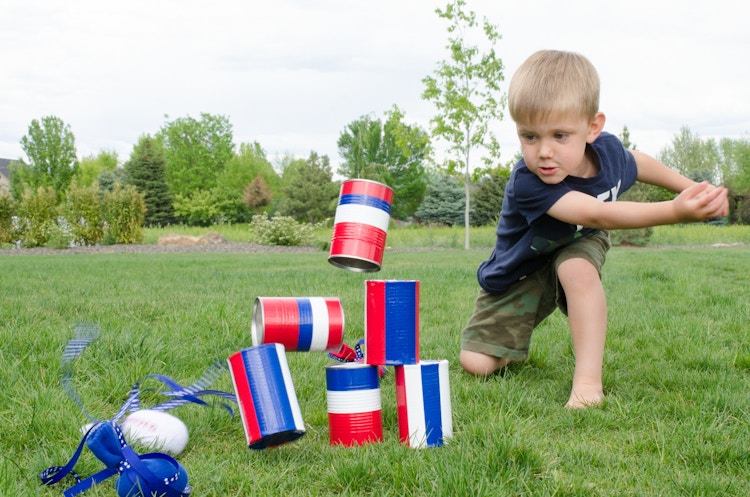 A child throwing a beanbag made of a sock filled with rice at a stack of red, white, and blue striped cans and knocking them down in the backyard.
