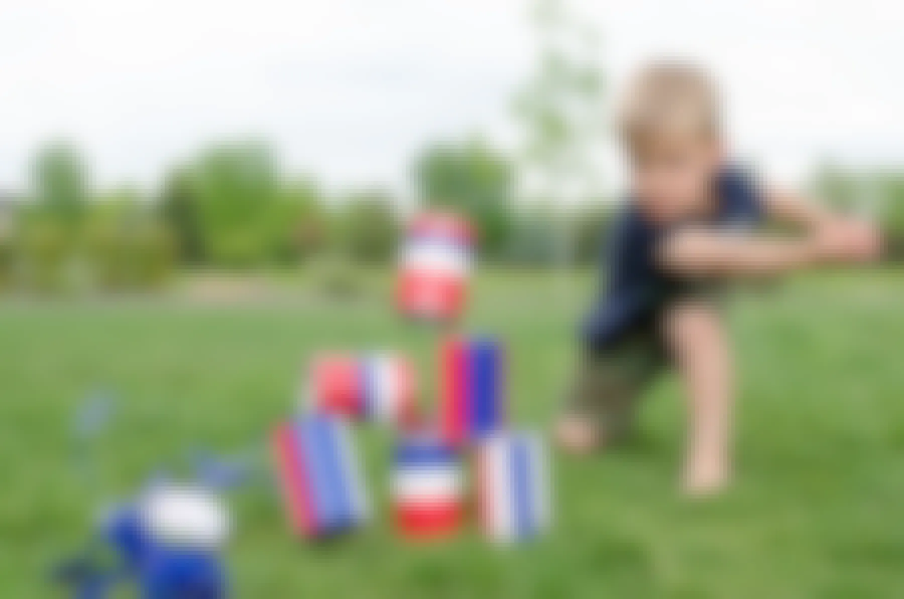 A child throwing a beanbag made of a sock filled with rice at a stack of red, white, and blue striped cans and knocking them down in the backyard.