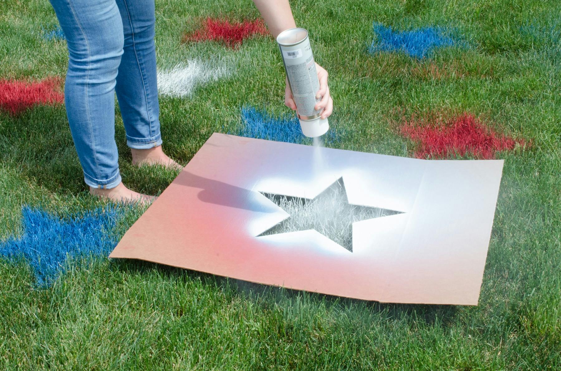 A person using a star-shaped cardboard stencil and marking spray paint to put red, white, and blue stars on a lawn.
