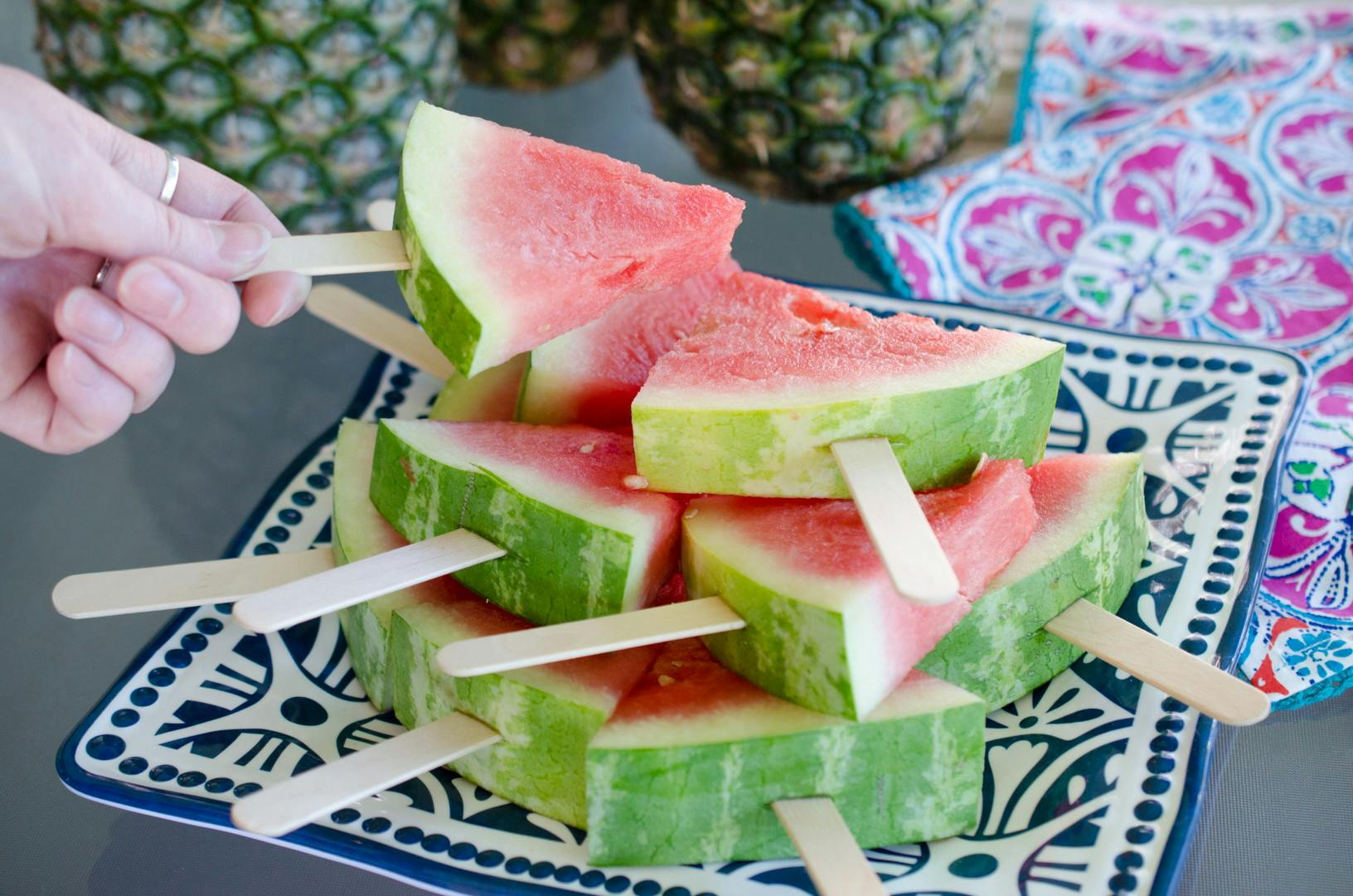 A person picking up a watermelon slice with a popsicle stick in it off of a plate of more of the same.