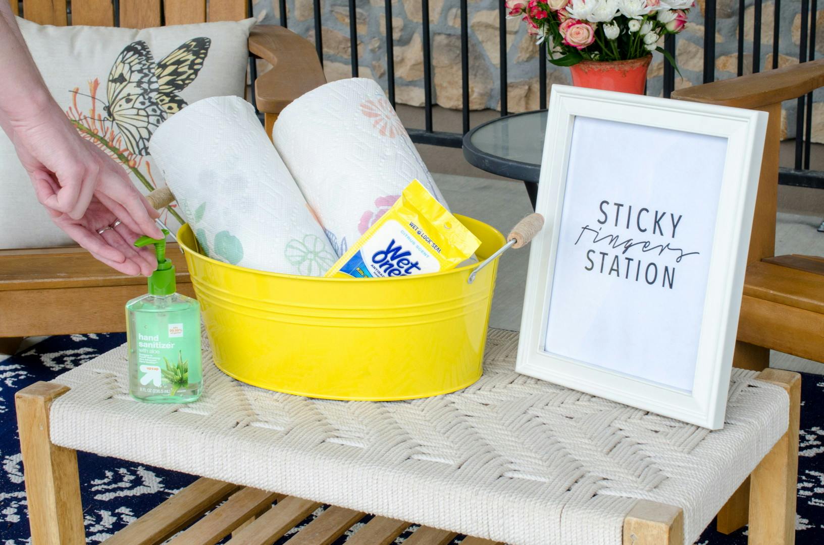 Person reaching for hand sanitizer next to a bucket with rolls of paper towels and Wet Ones on a table with a sign that reads, "Sticky fingers station.