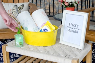 Person reaching for hand sanitizer next to a bucket with rolls of paper towels and Wet Ones on a table with a sign that reads, "Sticky fingers station.