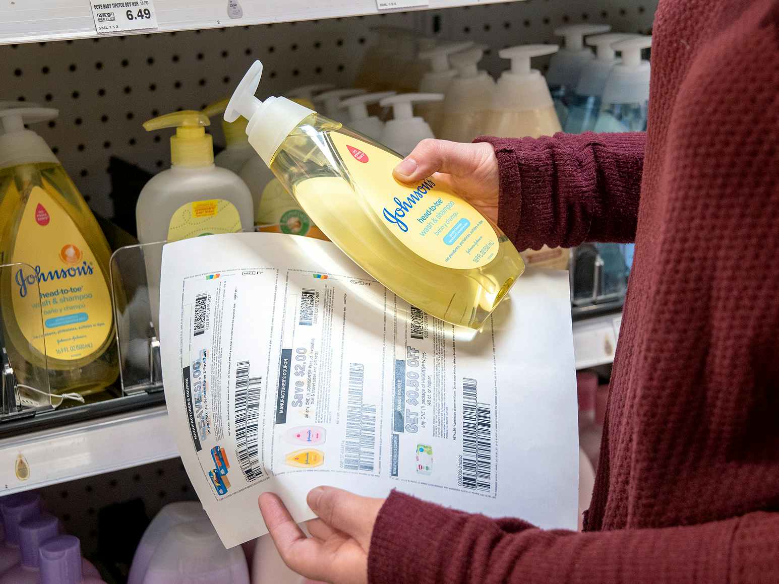 person holding a sheet of paper with coupons on it while holding a bottle of Johnson and Johnson baby shampoo in front of store shelves with baby shampoo on them.