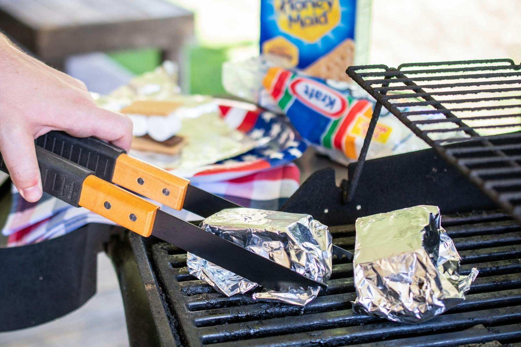 A person's hand using tongs to pick up a foil-wrapped s'more from a grill next to another foil-wrapped s'more, with s'more ingredients in the background.