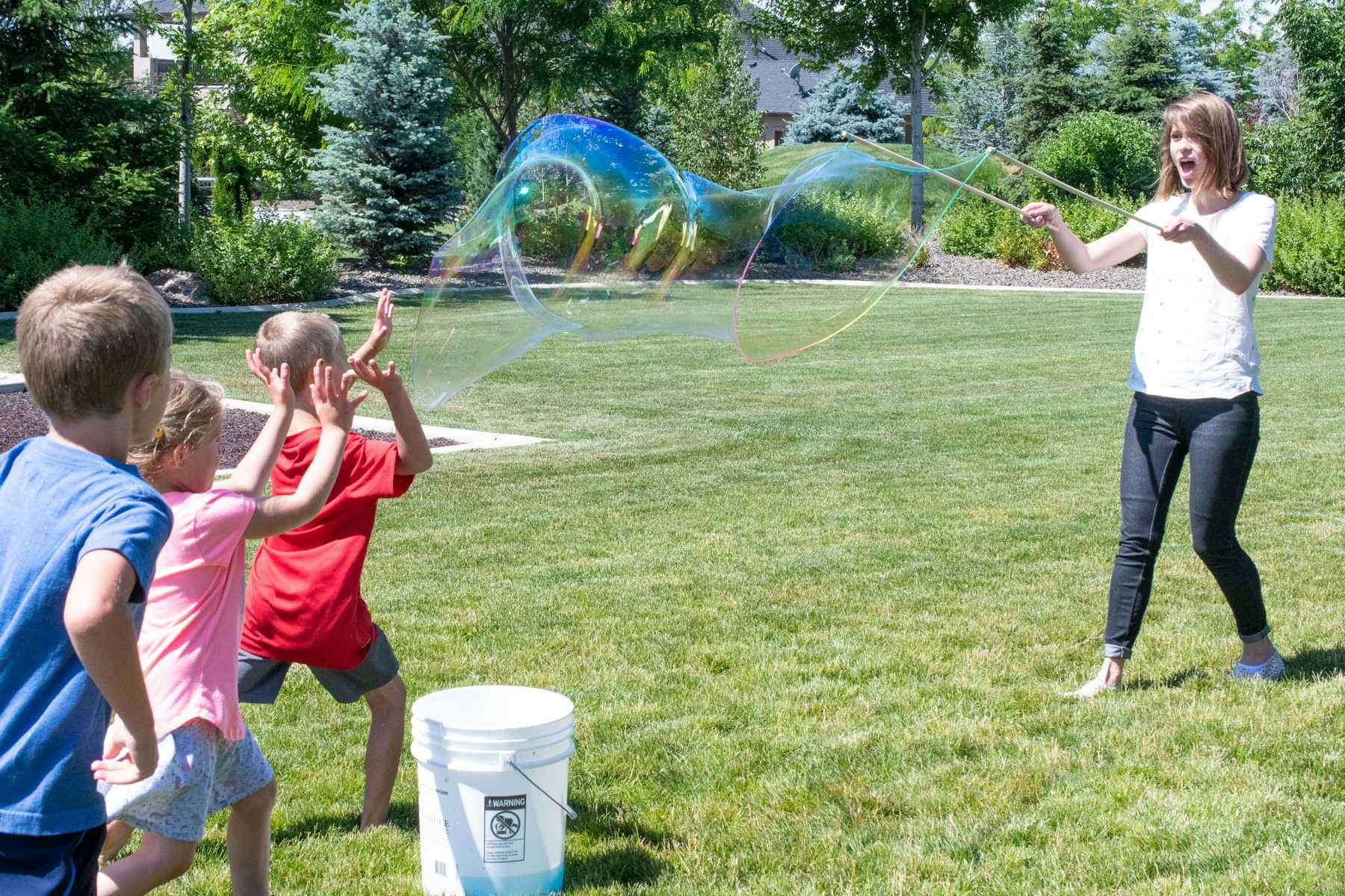 A woman holding two sticks to create a giant bubble in a backyard next to a bucket, and children running up to play.