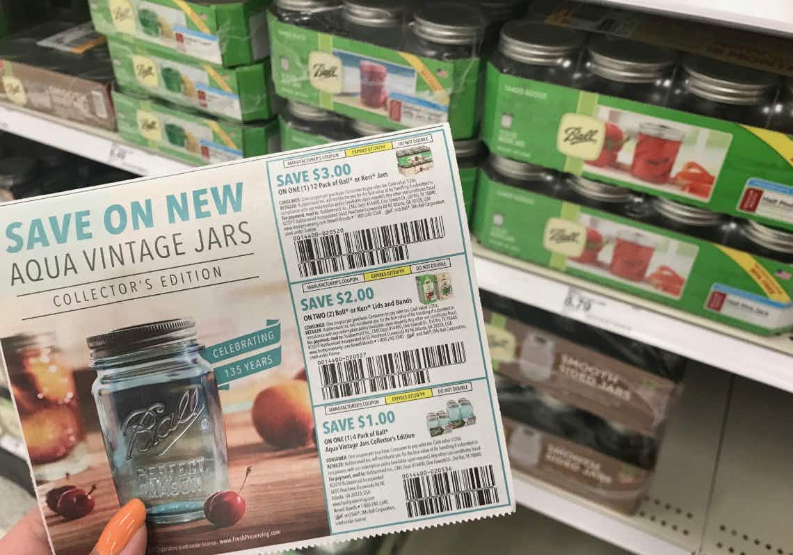 A person holding a coupon for Ball mason jars in front of a shelf of Ball jars.