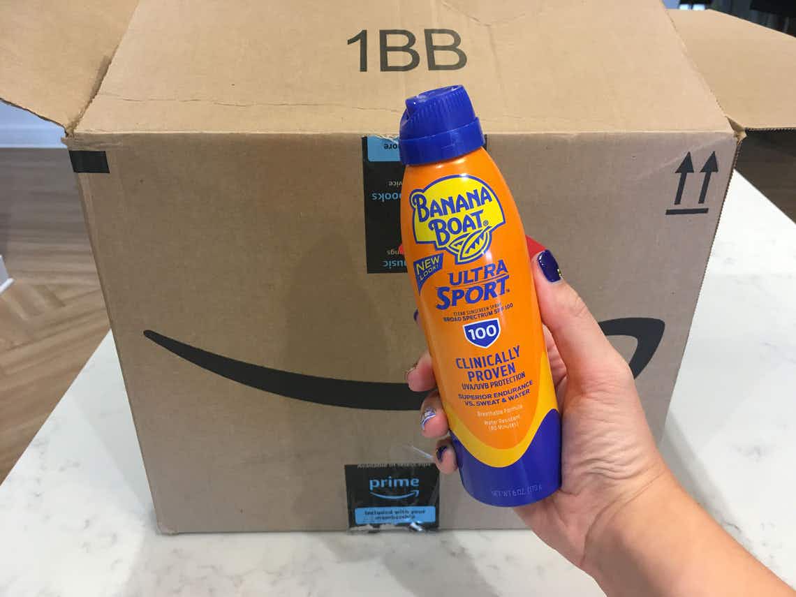 A hand holding a bottle of Banana Boat sunscreen in front of a delivery box.