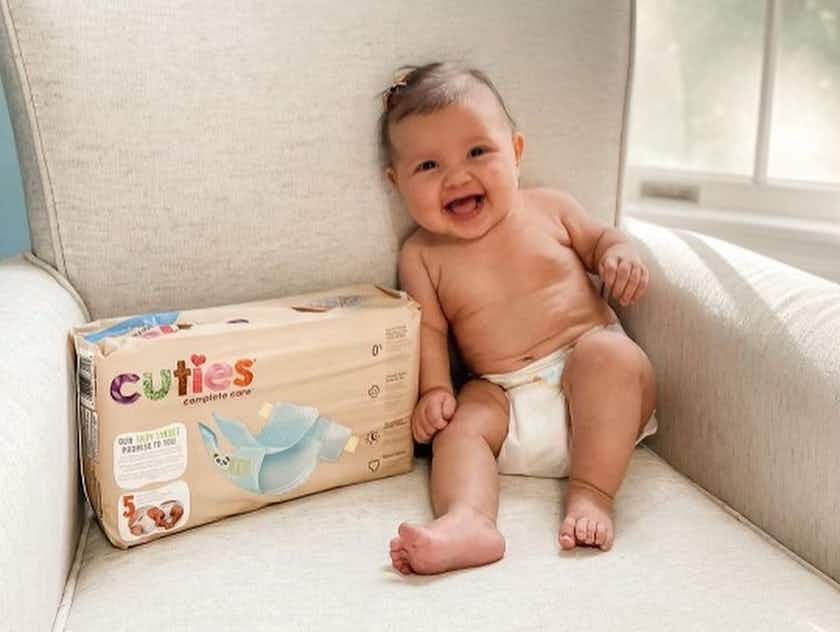A baby wearing only a diaper, sitting on a big beige chair next to a pack of Cuties baby diapers.