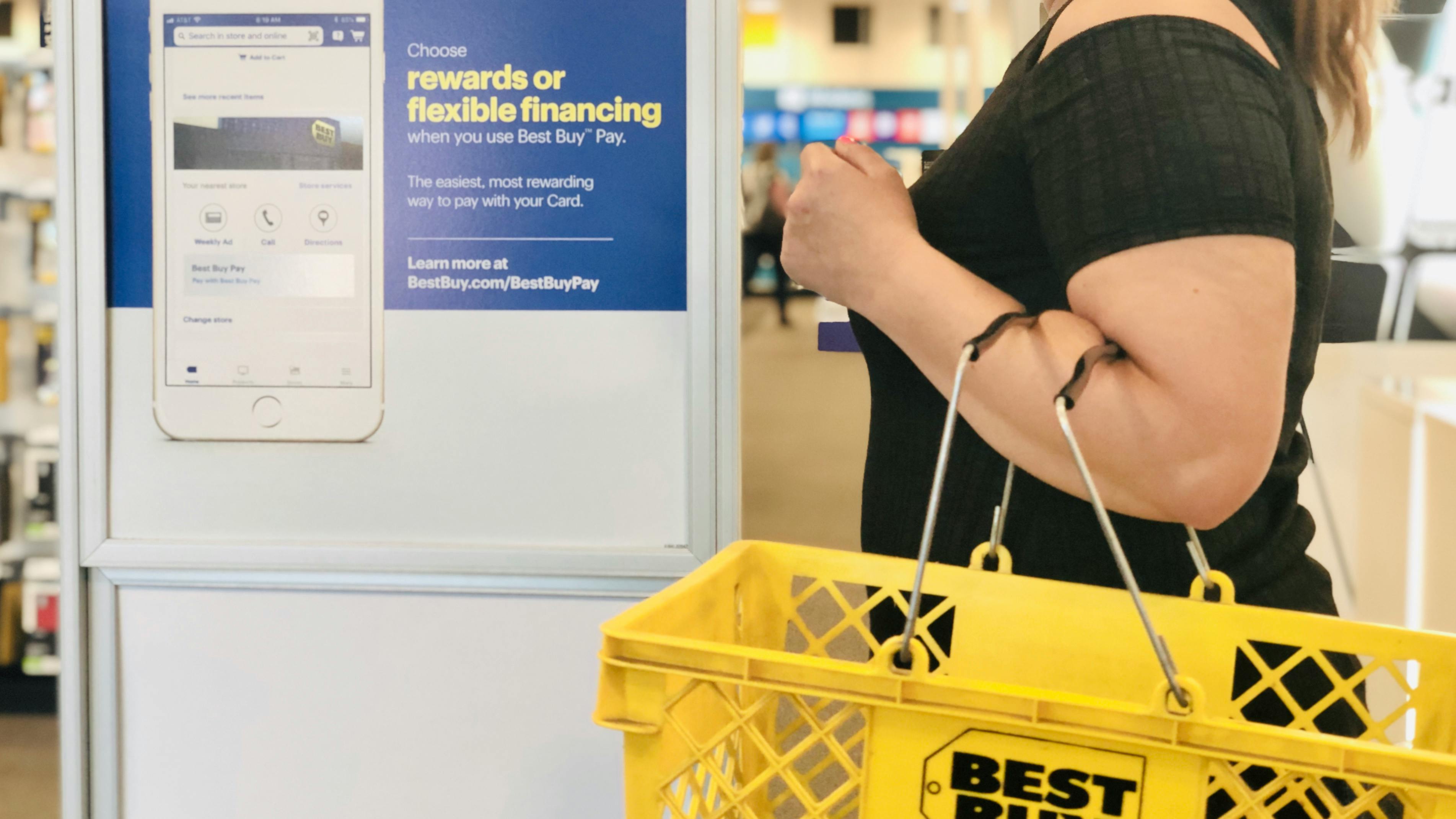 woman stands in front of a best buy rewards sign holding a yellow best buy shopping basket