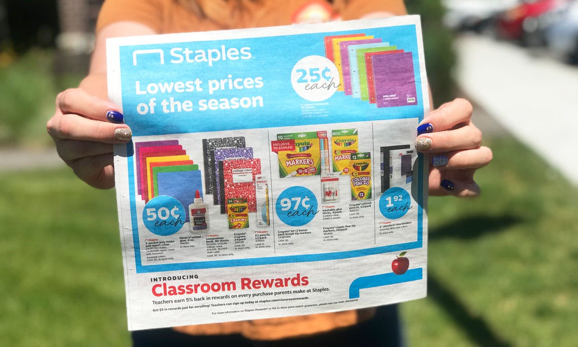 A person holding up a paper ad for Staples' Lowest prices of the season sales.