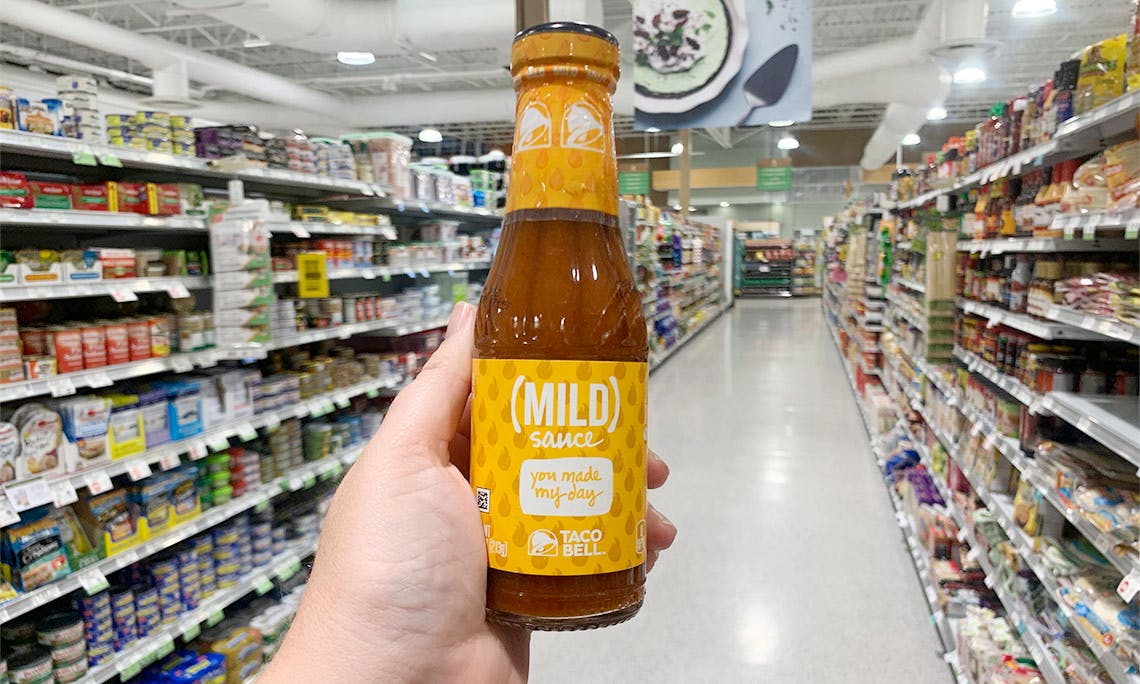 A person's hand holding up a bottle of Taco Bell Mild Sauce in a grocery store aisle.
