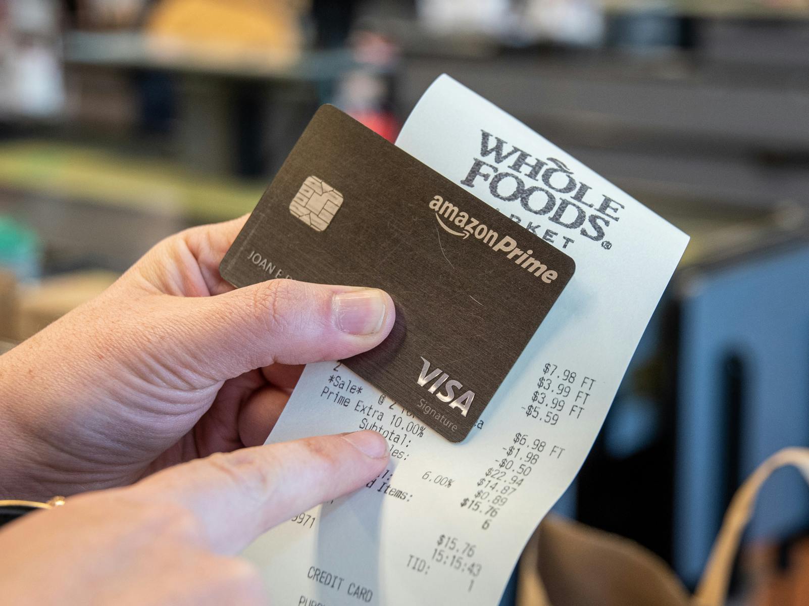 An Amazon Prime credit card being held up with a Whole Foods receipt. The person holding them is using their other hand to point at something on the receipt.