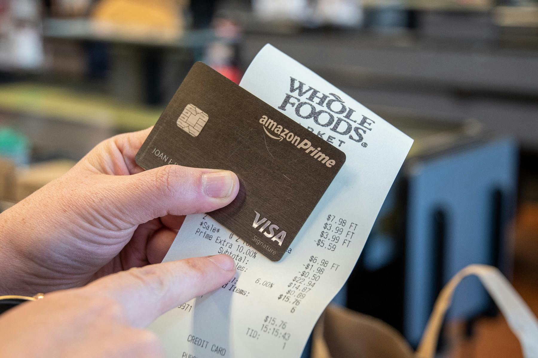 An Amazon Prime credit card being held up with a Whole Foods receipt. The person holding them is using their other hand to point at something on the receipt.