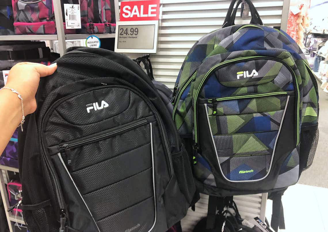 A person's hand holding up a FILA backpack in front of a sale display with more FILA backpacks and a SALE sign inside Kohl's.