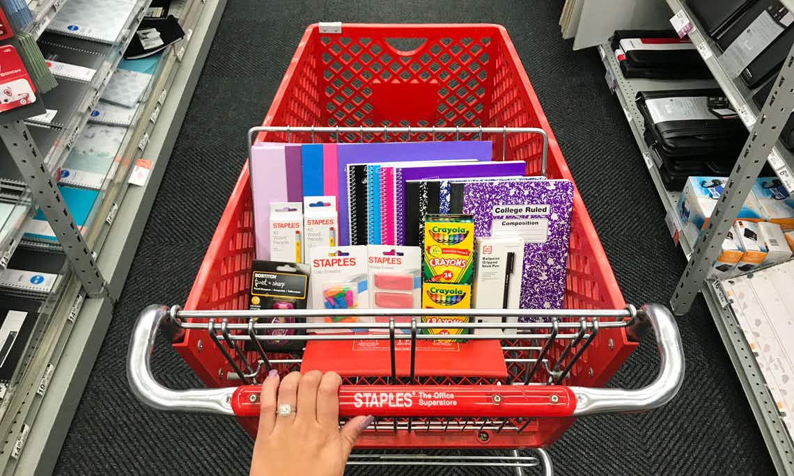 A person's hand pushing a Staples shopping cart with school supplies in it.
