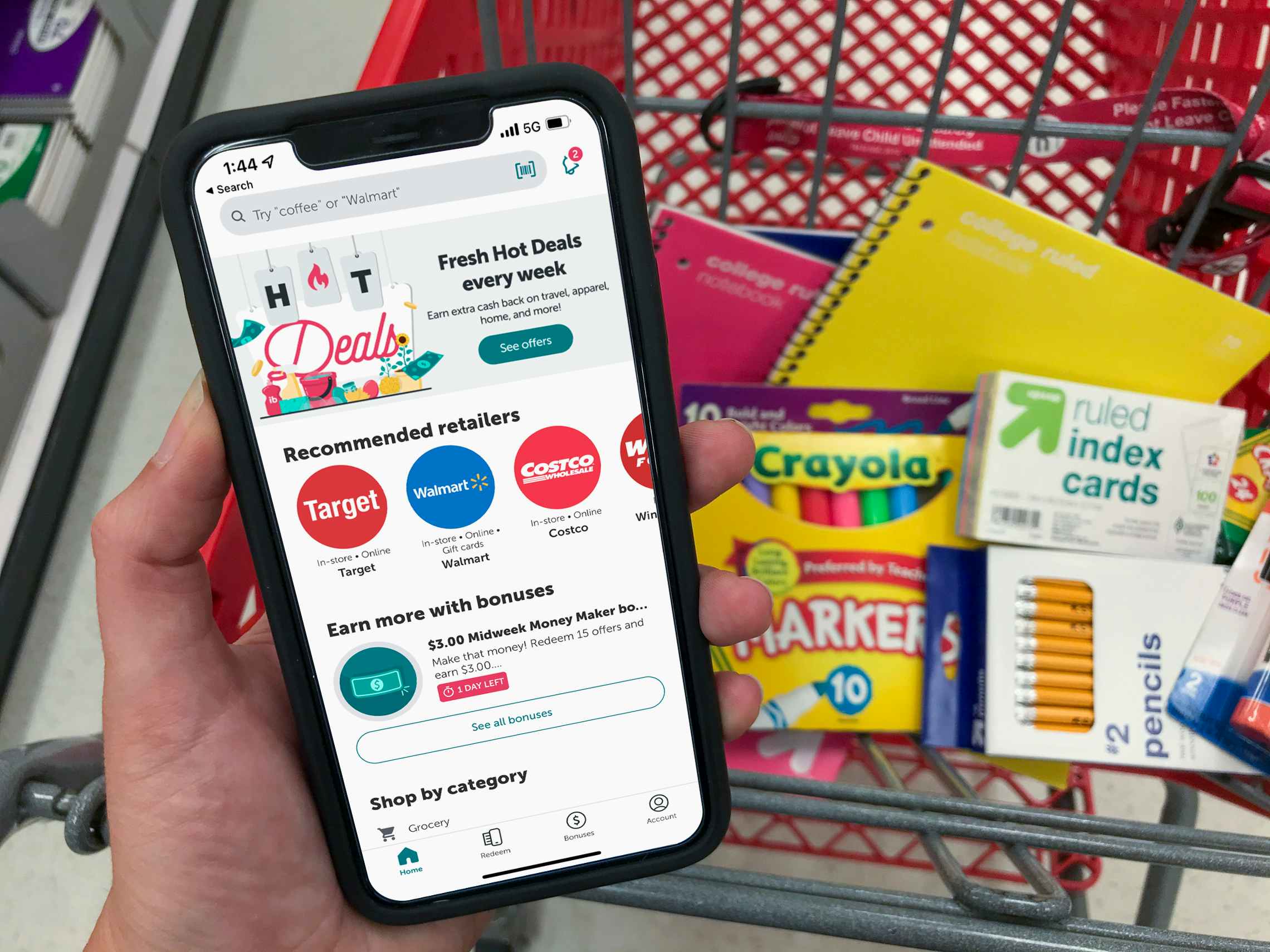 A person's hand holding a phone displaying the ibotta app main page above a shopping cart with the basket filled with school supplies.
