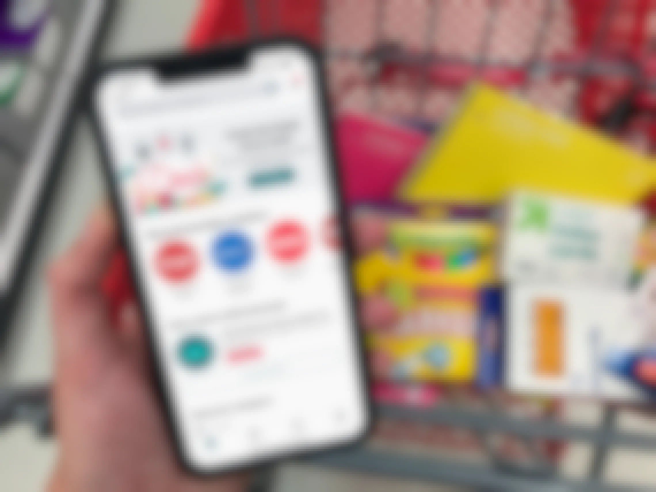 A person's hand holding a phone displaying the ibotta app main page above a shopping cart with the basket filled with school supplies.
