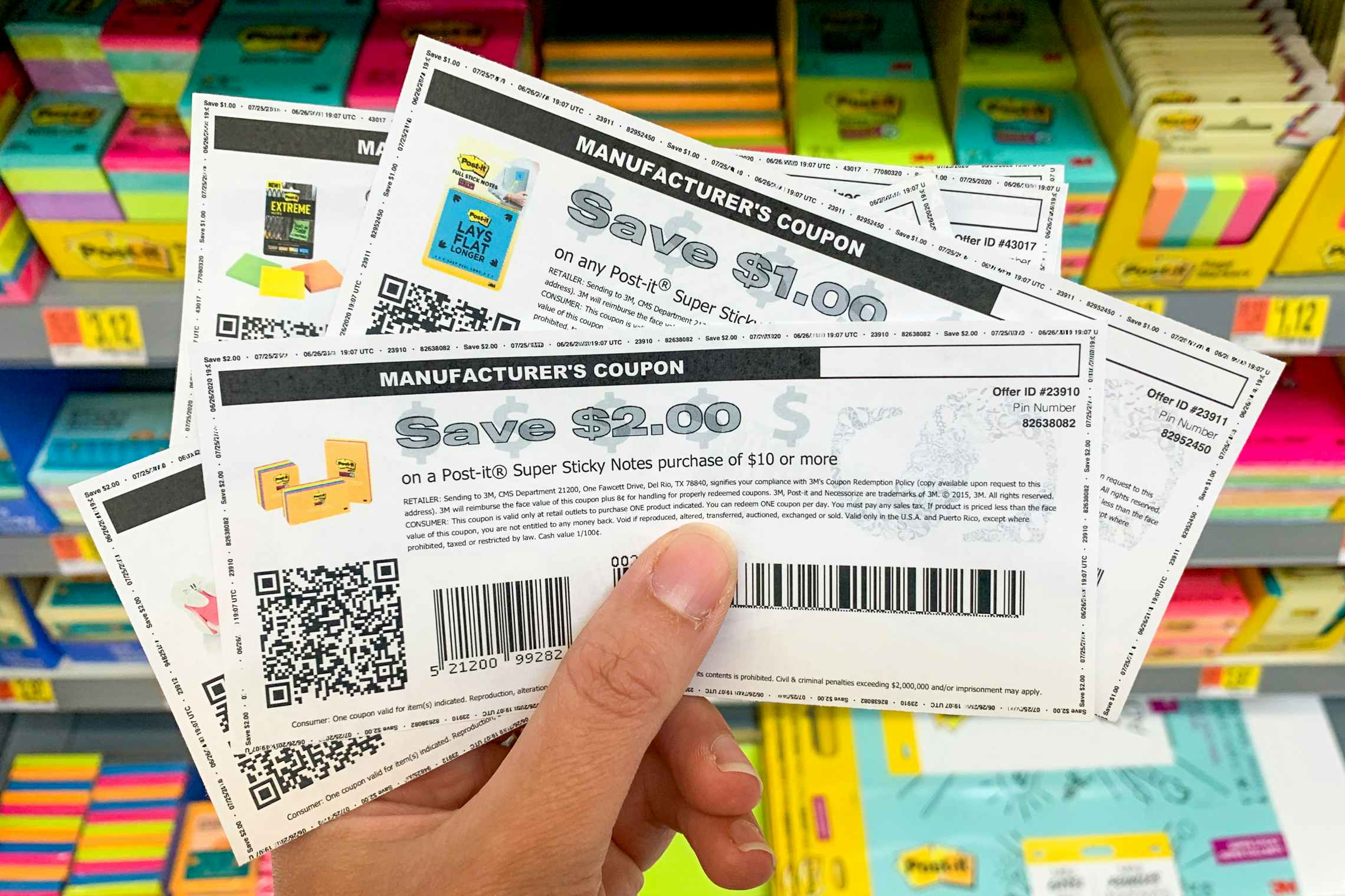 A person's hand holding some manufacturer's coupons for Post-it products in front of a shelf filled with post-it products.
