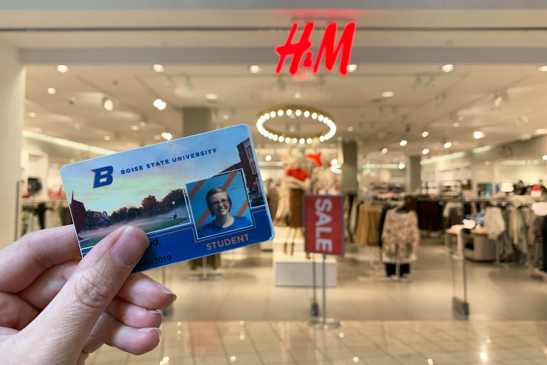 A person's hand holding a Boise State University student ID in front of an H&M storefront inside of a mall.