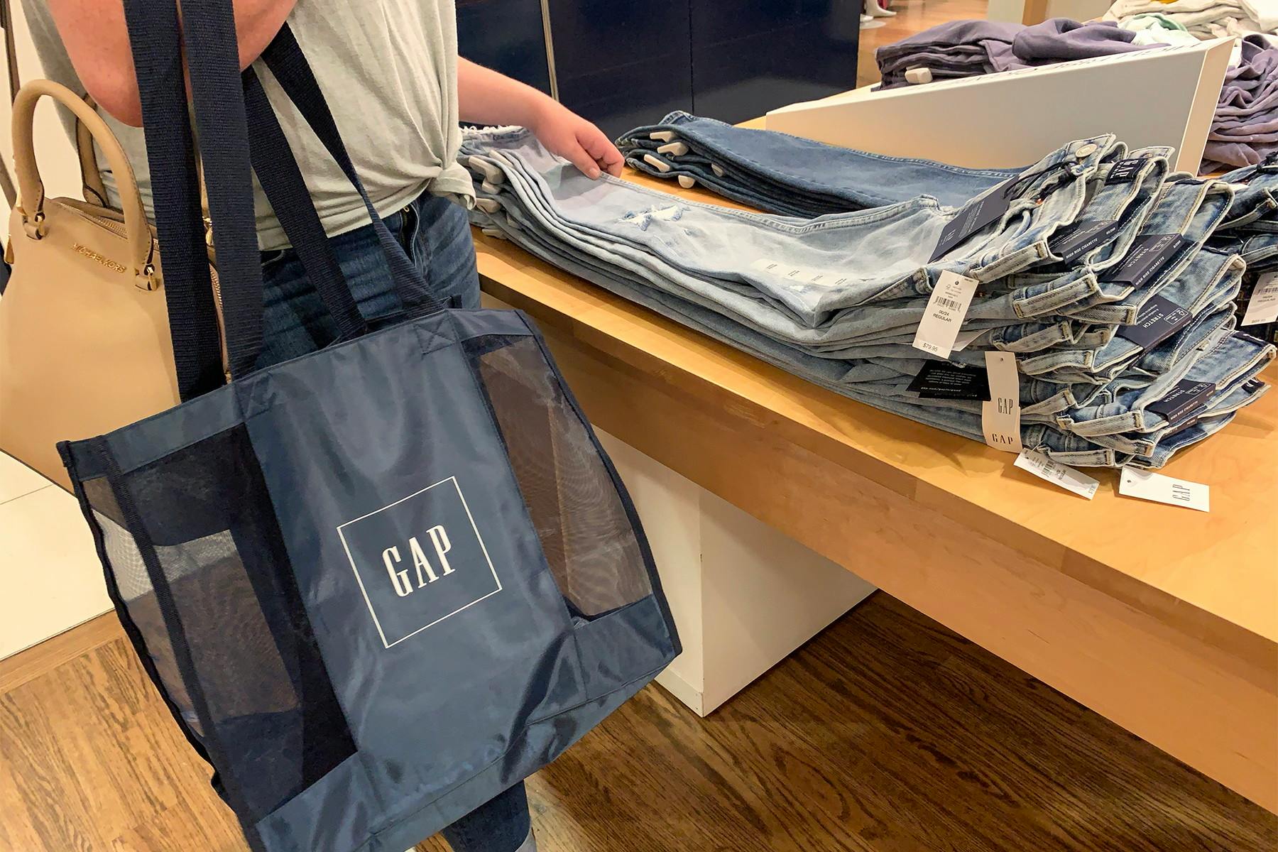 Someone carrying a GAP shopping bag and looking at jeans on a table in a GAP store