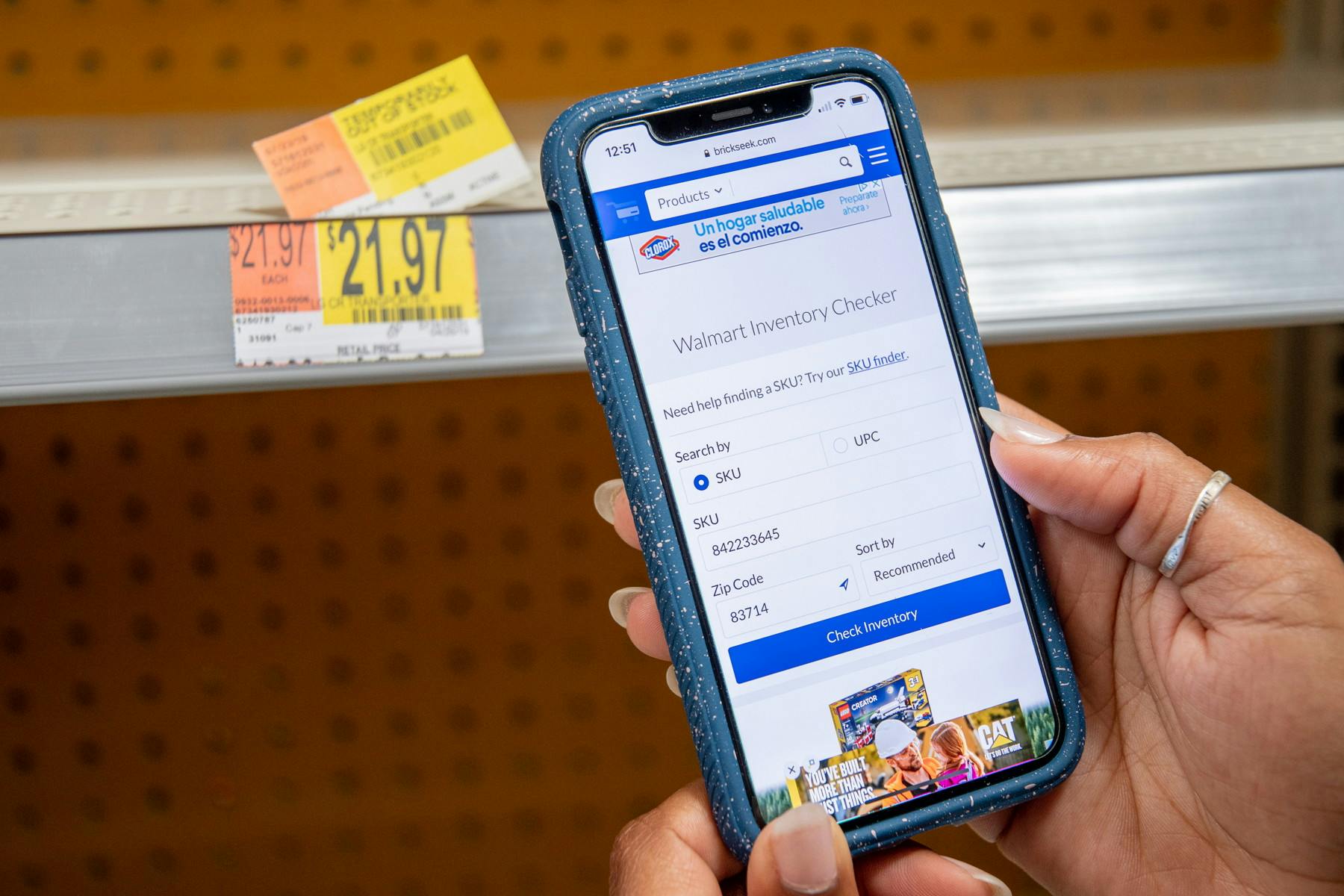 A person's hands holding a cell phone displaying BrickSeek.com's Walmart Inventory Checker in front of an empty shelf.