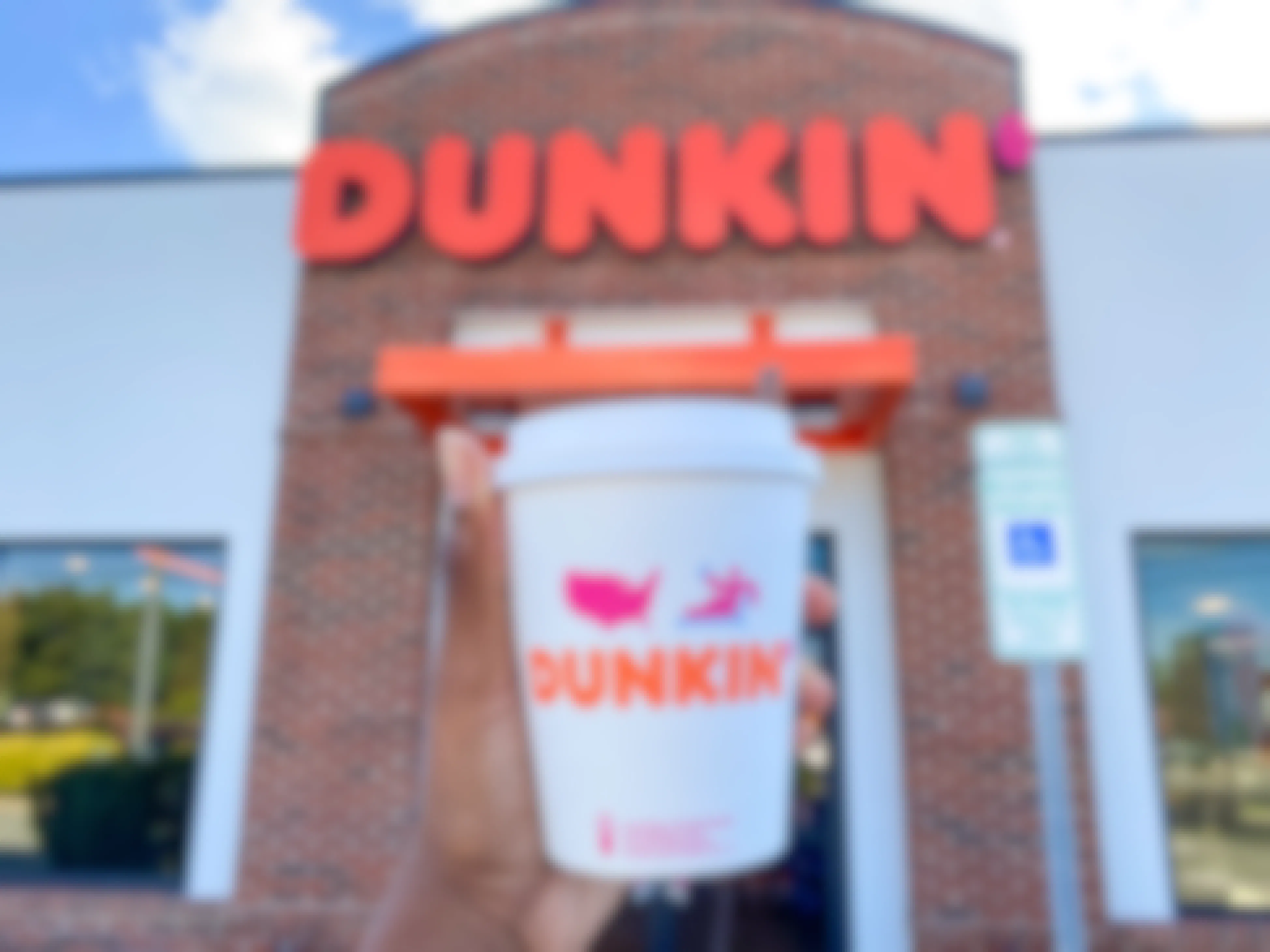 A person's hand holding up a Dunkin pumpkin spice latte in front of a Dunkin restaurant.