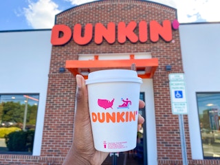 A person's hand holding up a Dunkin pumpkin spice latte in front of a Dunkin restaurant.