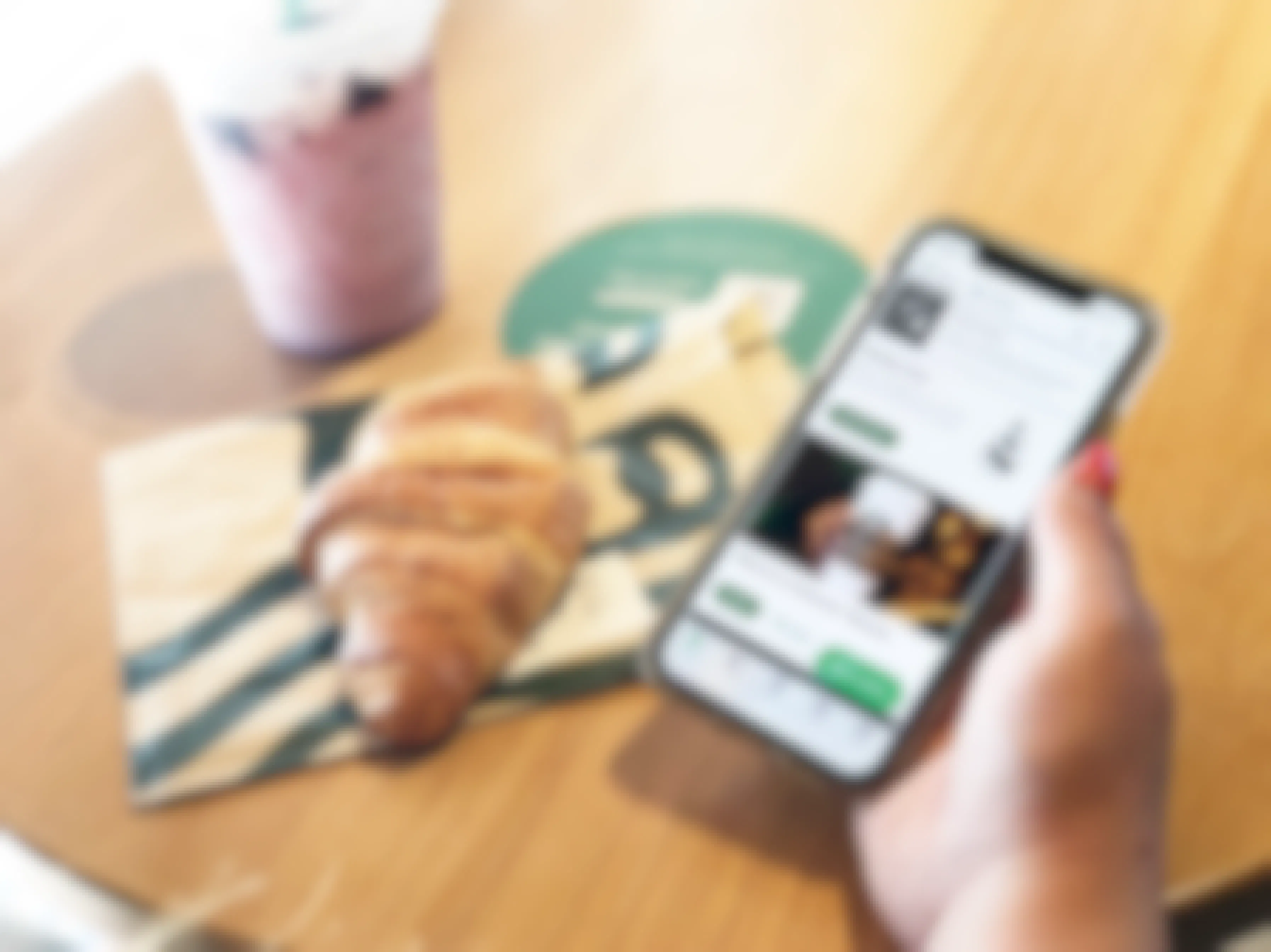 A person holding a smartphone with the Starbucks app open above a table with a croissant and drink sitting on it.