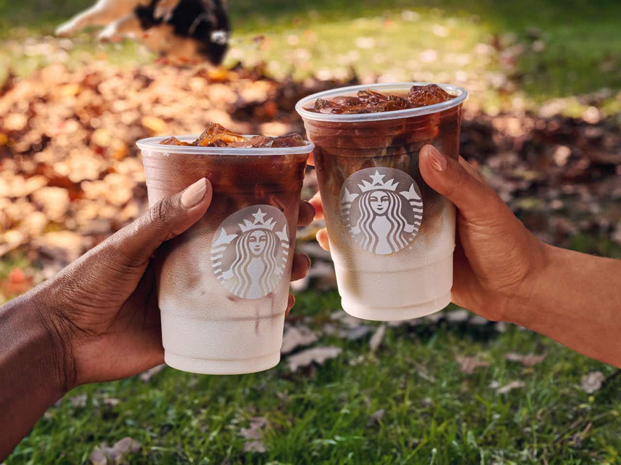 two peoples hands holding pumpkin cream cold brews that are being held by two people with autumn leaves in the background