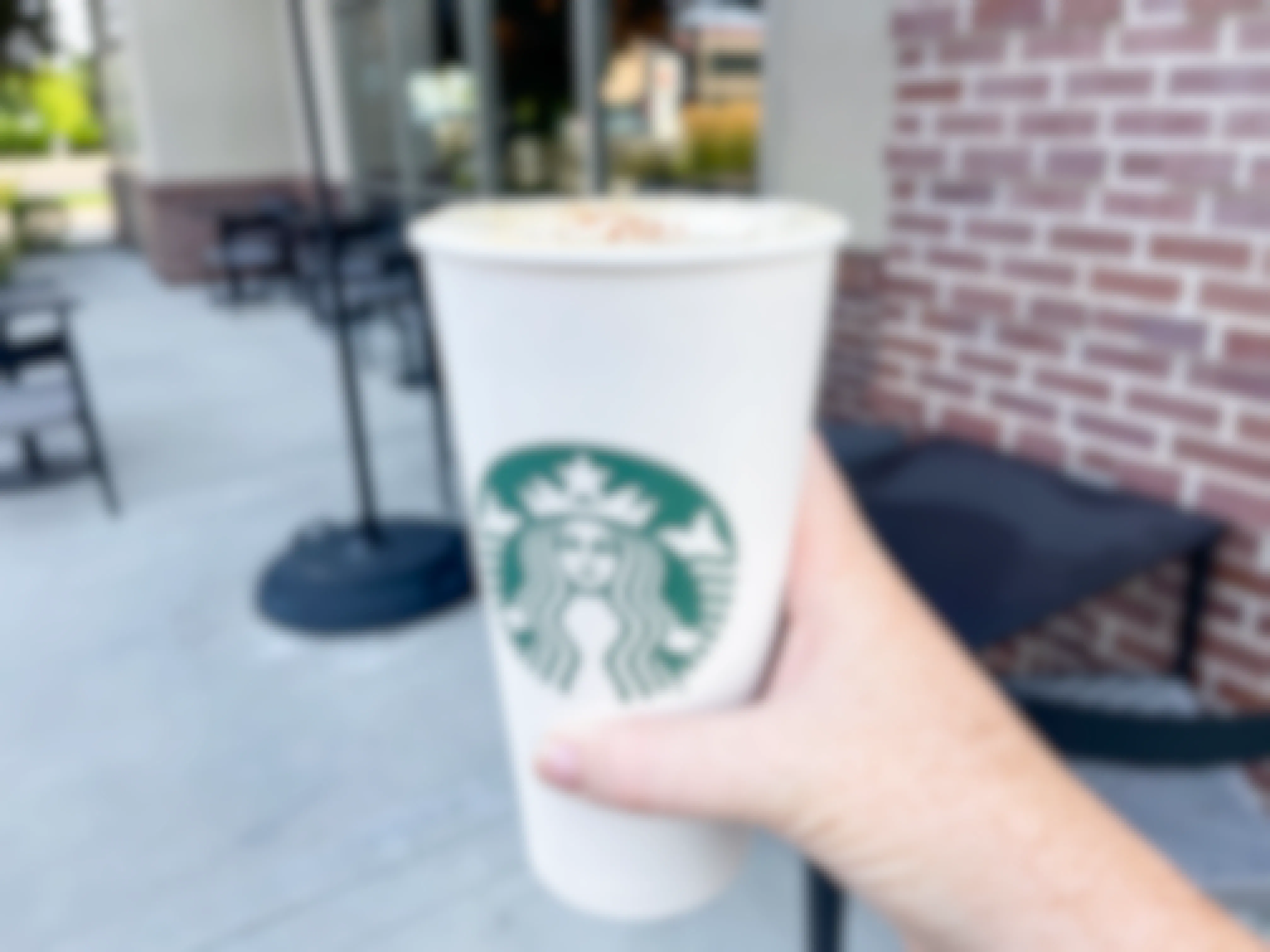 A person's hand holding a Starbucks pumpkin spice latte in front of a Starbucks.