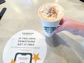 Someone picking up a Starbucks Pumpkin Spice Latte from a counter with rewards sticker on it.