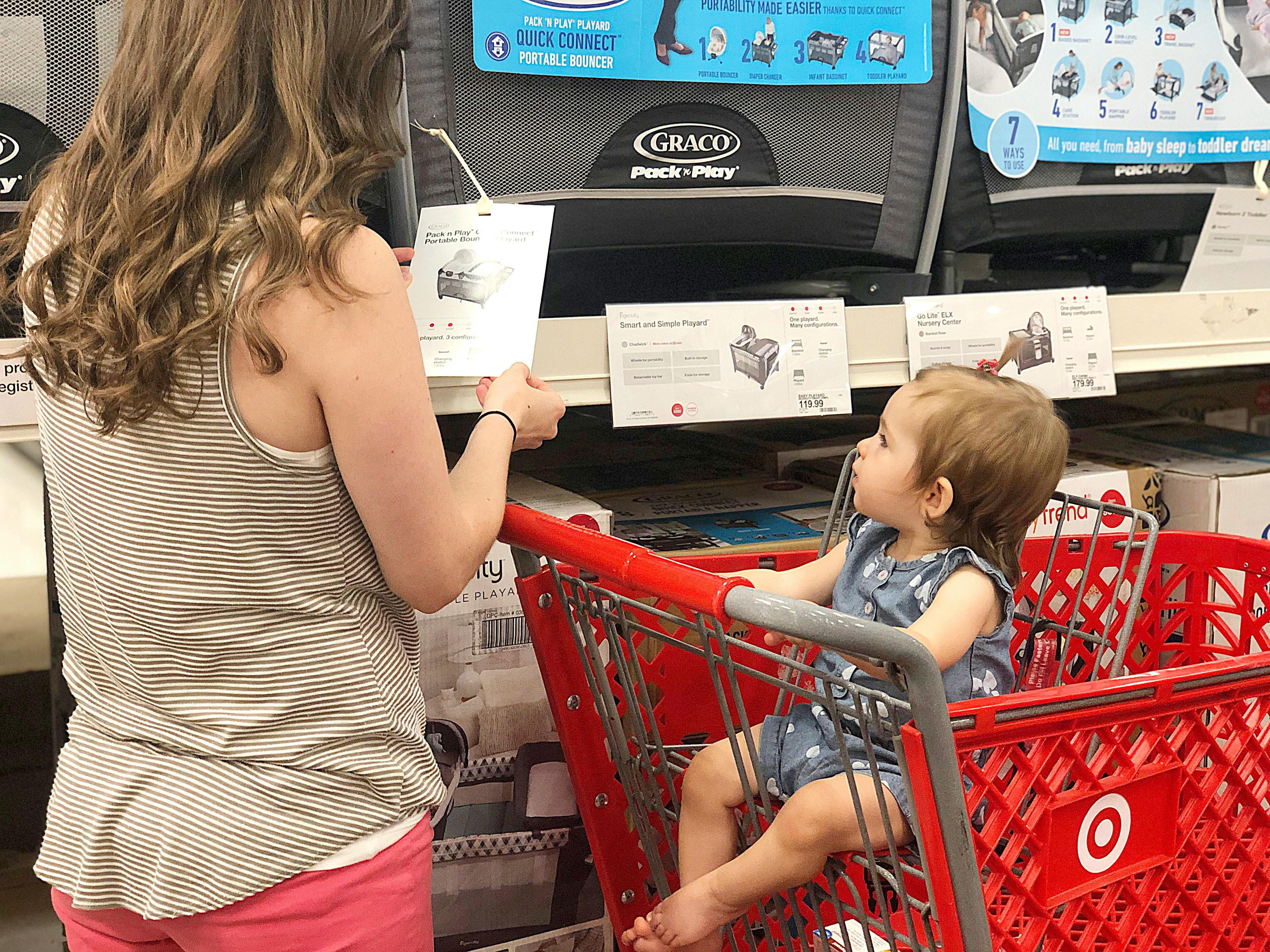 A mom looking at an information card about a crib next to her baby sitting in the basket of a Target shopping cart.