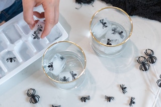 Someone taking ice cubes with plastic spiders in them out of an ice tray and putting them in glass drinkware.