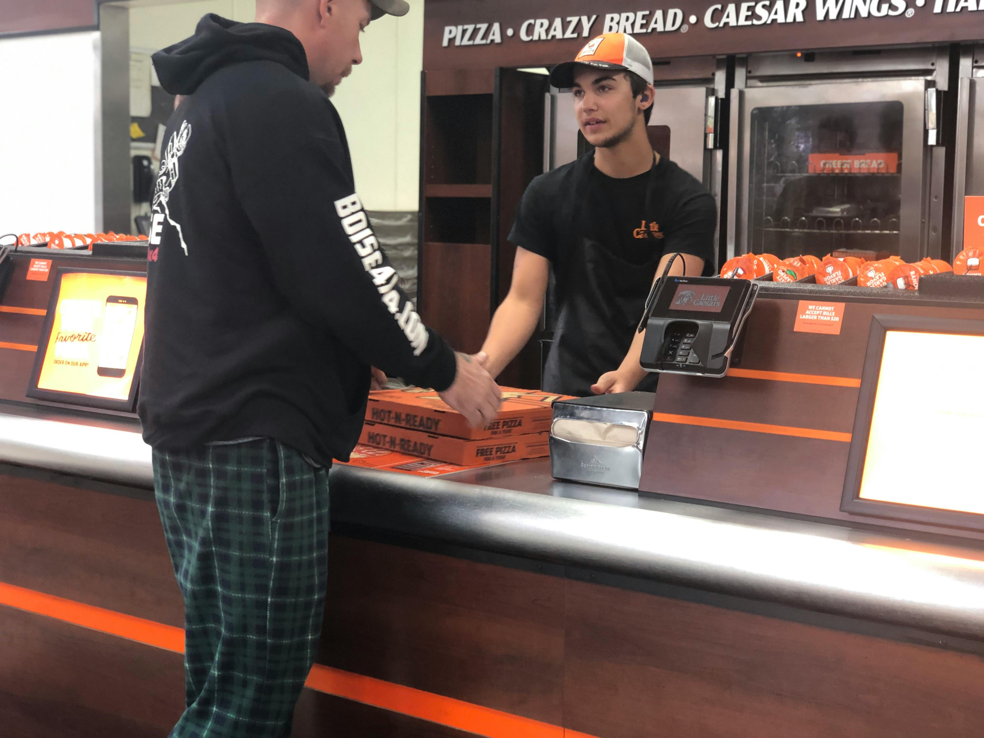 A Litlle Caesars employee behind a restaurant ocunter giving a person a pizz box.