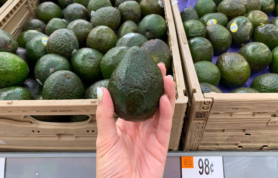 left hand holding greenish-black avocado in front of other avocados in tan crates at walmart