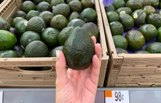 left hand holding greenish-black avocado in front of other avocados in tan crates at walmart