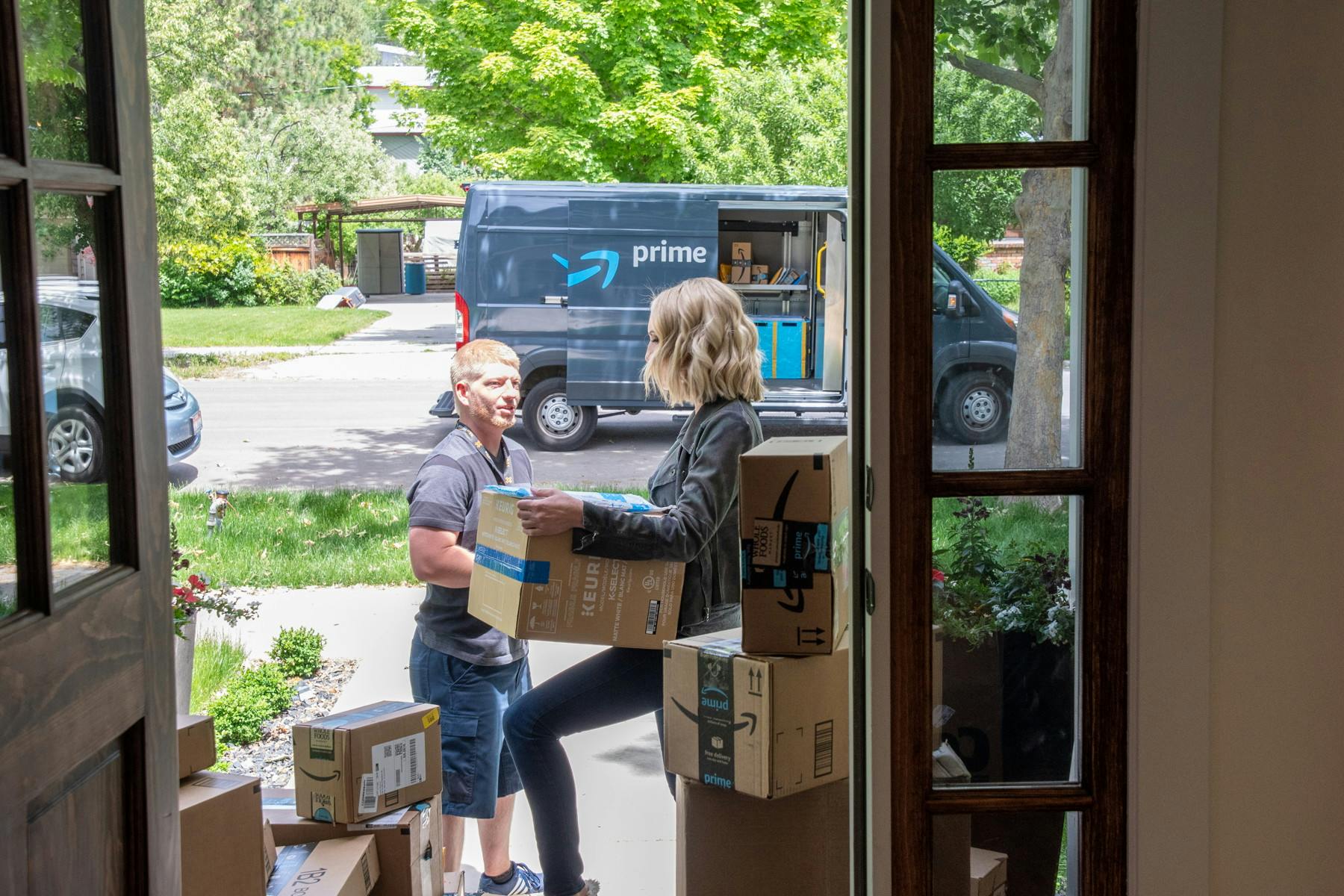 A woman holding a box and talking to an Amazon delivery driver on her front porch which is covered in Amazon packages.