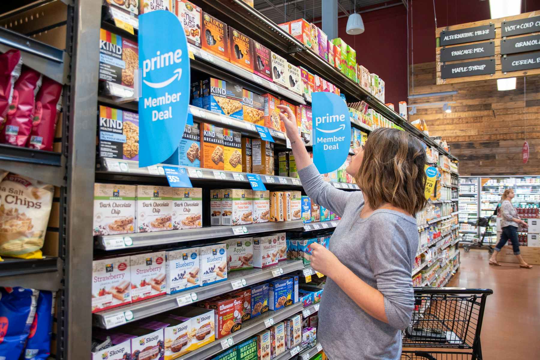 A woman shopping at Whole Foods, reaching up to a shelf tagged with blue Amazon Prime Members Deal signs.