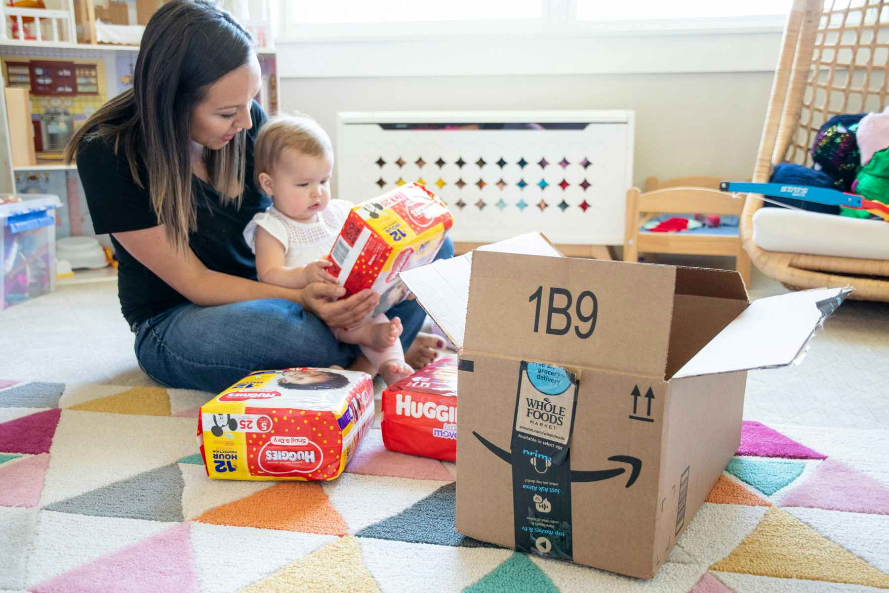 A woman and baby sitting on the floor of a nursery with packages of diapers and an amazon box