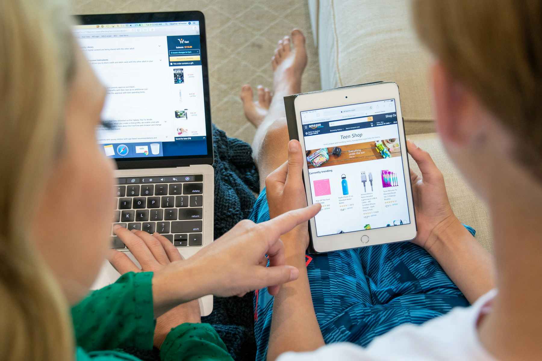 Two people sitting on a couch, one of them using a laptop and the other using a tablet, both of them looking at the Amazon website.