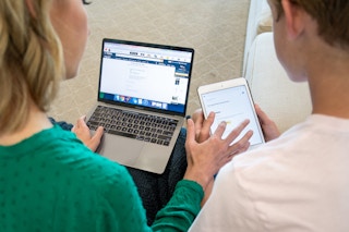 a woman using a a laptop displaying Amazon and clicking something on the tablet of the boy sitting next to her.