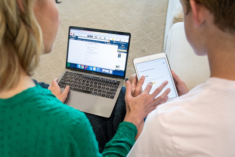a woman using a a laptop displaying Amazon and clicking something on the tablet of the boy sitting next to her.